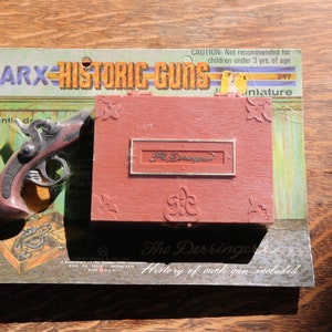 Op de afbeelding: Een vintage Marx Historic Guns doos met een rode en witte doos en een klein, zilverkleurig pistool. De doos heeft de tekst "The Derringer" en "History of each gun included".