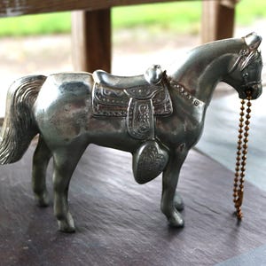 Può includere: Statuetta di cavallo color argento con sella e briglia. Il cavallo ha una lunga coda e una catena di perline che pende dalla bocca. La sella è decorata con motivi decorativi. La statuetta è su una superficie scura.