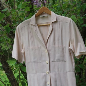May include: A light beige button-up dress with short sleeves and two patch pockets. The dress has a collared neckline and a button closure.