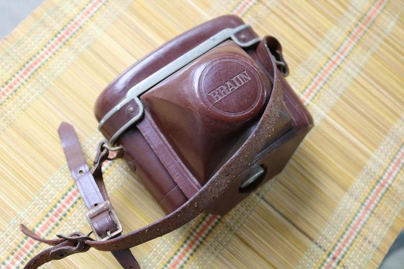 Pu&ograve; includere: Una custodia per fotocamera in pelle marrone con finiture in metallo argentato e una tracolla. La custodia ha la scritta "BRAUN" in rilievo sul davanti.