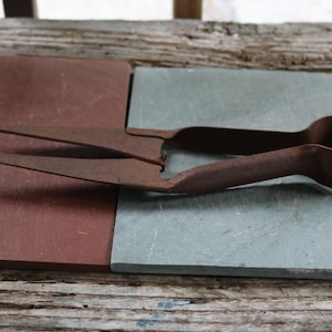 May include: A rusty, vintage metal grass shear tool with long blades and a looped handle. The shears are resting on a surface of red and gray slate tiles, with a weathered wooden background.