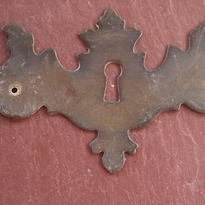 May include: Ornate antique brass keyhole cover with decorative scrollwork and a central keyhole. The cover has two small holes for mounting and is set against a reddish-brown surface.