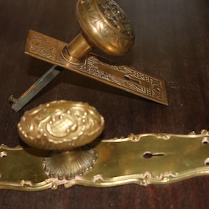 May include: Two brass door knobs with matching rectangular backplates. One knob is attached to a rectangular plate with an integrated keyhole and a metal spindle. The second knob features an oval shape and sits on a decorative, scalloped-edge plate with a keyhole and screw holes.