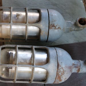 Op de afbeelding: Twee vintage industriële lampen. Elke lamp heeft een cilindrische glazen behuizing beschermd door een metalen kooi. De lampen zijn grijs geschilderd met zichtbare roest en hebben een gebogen pijp die vanaf de zijkant uitsteekt.