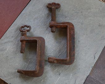 May include: Two antique C-clamps made of rusty metal, with threaded screws and handles. One clamp has a clover-shaped handle, the other a T-shaped handle. The clamps are on a gray and brown surface.