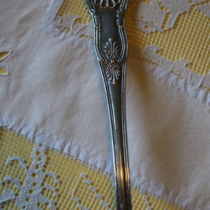May include: A silver-colored utensil with an ornate handle. The handle features a detailed design with a shell-like top and a central floral motif. The utensil is set against a yellow and white patterned background, suggesting a vintage or antique aesthetic.