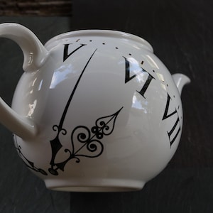May include: White ceramic teapot with a black clock face design. The teapot features a curved handle and a spout. The clock design includes Roman numerals and stylized clock hands. A decorative kitchenware item.
