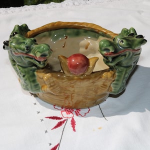 Könnte beinhalten: Eine Keramikschale mit Drachendesign. Die Schale ist grün, beige und rot, mit zwei Drachenköpfen auf jeder Seite. In der Mitte befindet sich eine rote Kugel. Die Schale hat einen geflochtenen Griff.