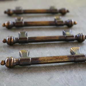 May include: Four metal drawer pulls with a dark, aged brass finish. Each handle features a central cylindrical bar, two mounting posts, and decorative rounded finials at both ends, arranged in a row from the foreground to the background.