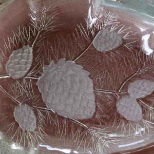 May include: Clear glass serving dish with a frosted pine cone and branch design. The dish has a leaf shape with a small handle. The design is etched into the glass, creating a textured effect.