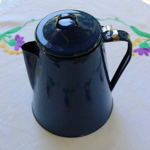 Può includere: Una caffettiera in smalto blu scuro con coperchio e manico nero. La caffettiera ha un beccuccio per versare e un fermaglio in metallo color argento sul manico. È appoggiata su una tovaglia bianca con fiori ricamati.