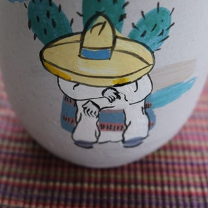 May include: A white ceramic vase featuring a hand-painted cartoon figure in a yellow sombrero and a blue and white striped serape. The design includes a green cactus and blue accents.