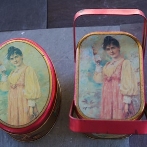May include: Two vintage red and gold metal containers. One is oval-shaped, the other rectangular with a handle. Both feature a woman in a pink and yellow dress holding a bottle. The containers have a vintage aesthetic.
