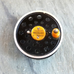 May include: A black Pflueger Medalist fishing reel with a yellow metal spool and a black knob. The reel has a white rim and a yellow label that reads "Pflueger Medalist by Shakespeare".