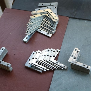 Peut inclure: Plusieurs équerres métalliques argentées en forme de L, avec des trous pré-percés, disposées sur une surface brun-rouge et grise. Idéales pour la construction et l'assemblage de meubles.