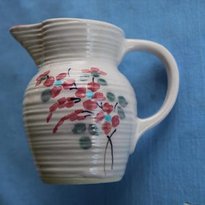 Puede incluir: Una jarra de cerámica blanca con asa y pico. La jarra tiene una textura acanalada y está decorada con diseños florales rojos, azules y verdes. El fondo es una tela azul.