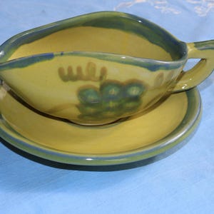 May include: A yellow ceramic gravy boat and saucer set. The boat has a handle and a curved spout, with a blue-green rim and grape design. The saucer is round with a matching rim. The set is on a blue surface.