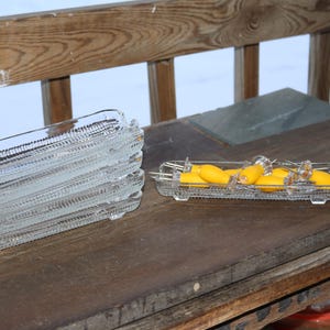 May include: A stack of clear, rectangular glass serving dishes with a textured design. Another dish holds yellow fruit, possibly lemons, with clear glass serving utensils. The dishes are on a wooden surface.