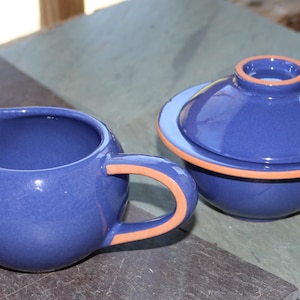May include: A blue ceramic creamer with an orange handle and a matching blue ceramic bowl with a lid. The creamer and bowl are on a slate-colored surface. The creamer has a rounded shape and a spout.