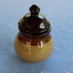 Pode incluir: Um pequeno pote de cerâmica redondo com tampa. O pote é bege com uma faixa marrom no meio e uma tampa marrom escura. A tampa tem um pequeno botão pontiagudo na parte superior. O pote está sobre uma superfície azul.