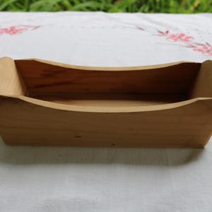May include: A small, rectangular wooden box with rounded ends. The box is made of light-colored wood and has a simple, natural design. The interior is visible, showing the depth of the box. The box is sitting on a white surface with pink floral embroidery.