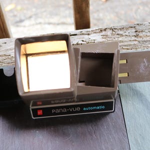 Puede incluir: Un proyector de diapositivas automático Pana-Vue vintage de color marrón y negro. El proyector tiene una ventana de visualización de plástico transparente y una bandeja para diapositivas.