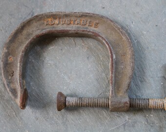 May include: An antique, adjustable C-clamp made of weathered metal. The word "ADJUSTABLE" is embossed on the top arch. The clamp has a threaded screw for adjusting the grip and is designed for woodworking or metalworking projects.