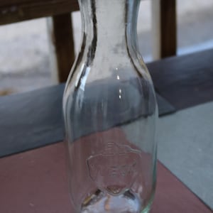 May include: Clear glass Weck jar with a wide mouth and a rounded body. The jar has a clear glass lid and a Weck logo embossed on the front. The jar is sitting on a brown surface.