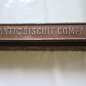May include: A rectangular metal display case with a weathered brown finish and a glass top. The front frame features the embossed text ATLANTIC BISCUIT COMPANY in raised silver-toned lettering. The metal surface shows signs of oxidation and wear.