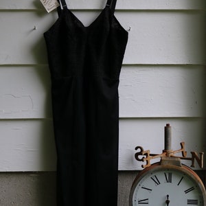 Op de afbeelding: Een zwarte kanten slipjurk hangt aan een witte hanger tegen een witte houten paneel achtergrond. De jurk heeft dunne bandjes en een kanten rand aan de onderkant. Een vintage-stijl klok is zichtbaar in de rechter benedenhoek van de afbeelding.