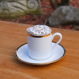 Könnte beinhalten: Eine weiße Porzellan-Teetasse mit goldfarbenem Rand und einem floralen Stoffdeckel. Die Teetasse steht auf einer weißen Untertasse mit goldfarbenem Rand.