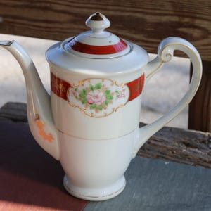 May include: White porcelain teapot with a red and gold floral design. The teapot has a lid with a knob and a curved handle.