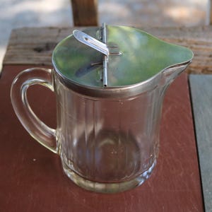 Könnte beinhalten: Ein klarer Sirupkrug aus Glas mit Griff und einem silberfarbenen Metalldeckel. Der Krug hat vertikale Rillen und einen Ausguss zum einfachen Ausgießen. Der Deckel hat ein Scharnierdesign.