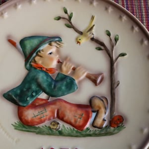 May include: A white ceramic plate with a raised image of a boy playing a flute. The boy is wearing a green hat, a green jacket, and red pants. A yellow bird is perched on a branch of a tree. The plate has a white background with a brown border. The plate is decorated with stars.