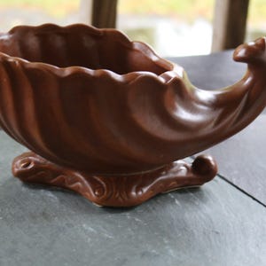 May include: A brown ceramic bowl with a scalloped rim and a curved, horn-like extension on one side. The bowl features a glossy finish and rests on a decorative, molded base with swirling patterns, presenting a distinct shell-like silhouette.