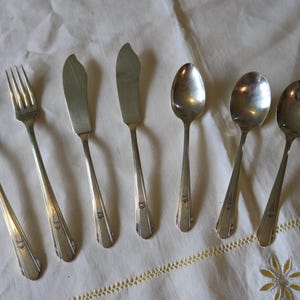 Op de afbeelding: Een set vintage zilveren bestek, inclusief vorken, botermessen en lepels, gerangschikt op een wit tafelkleed. Het bestek heeft een zilveren afwerking en een decoratief ontwerp op de handvatten. De afbeelding toont een klassieke eetservies.