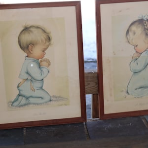 May include: Two framed vintage prints. Each print features a child kneeling in prayer, dressed in light blue clothing. The artwork has a soft, pastel color palette and is set against a light background. The frames are a dark brown color.