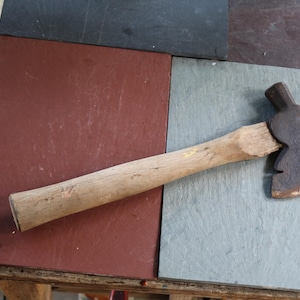 May include: A small hatchet with a weathered, light-coloured wooden handle and a dark, oxidised metal head. The metal head features a sharp, curved blade edge and a flat hammer poll on the opposite side, showing signs of age and heavy use.