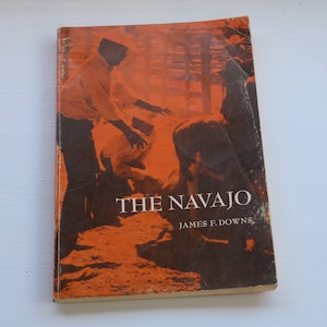 May include: A vintage book titled "The Navajo" by James F. Downs. The cover features an orange and brown illustration of people. The book has visible wear and tear.