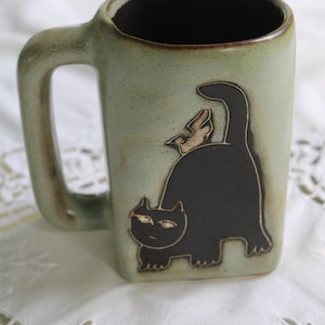 May include: A light green ceramic mug with a dark brown handle and interior. The exterior features a dark brown graphic of a cat with an arched back and a small bird perched on its tail. The mug has a square-like shape with rounded corners.
