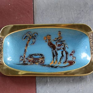 May include: Rectangular brass tray with a light blue enamel interior featuring a camel and palm tree design. The tray has decorative handles with green stone accents and coin-like embellishments. The tray is on a two-tone surface.