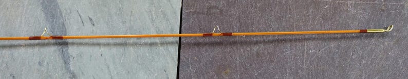 K&ouml;nnte beinhalten: Eine Angelrute aus Holz mit brauner und goldener Oberfl&auml;che. Die Rute ist etwa 1,8 Meter lang und hat eine goldene Spitze.
