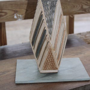 May include: A geometric ceramic vase with a diamond shape, featuring alternating sections of beige, brown, and gray. The vase is placed on a rectangular gray stone, showcasing a unique design. The vase is approximately 10 inches tall.