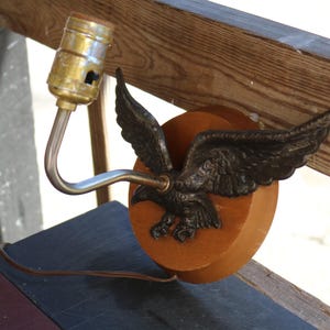 May include: Vintage wall sconce featuring a bronze eagle with outstretched wings, mounted on a round wooden base. The lamp has a brass bulb socket and a curved metal arm. A brown electrical cord extends from the base.