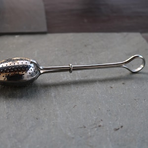 Può includere: Infusore da tè argentato con una ciotola perforata di forma ovale e un lungo manico con un anello all'estremità. Il filtro da tè è progettato per l'infusione di tè sfuso. L'infusore è in metallo lucido e poggia su una superficie grigia.