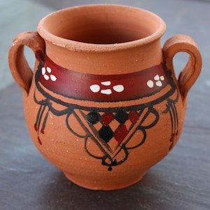 Puede incluir: Pequeño cuenco de cerámica color terracota con dos asas. El cuenco está decorado con una banda roja con puntos blancos y patrones geométricos negros y rojos. El diseño está pintado a mano, lo que le da un aspecto rústico y artesanal.