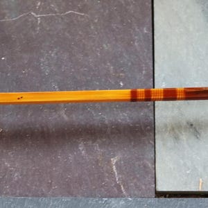 K&ouml;nnte beinhalten: Eine Angelrute aus Holz mit braunem und orangefarbenem Streifenmuster. Die Rute hat einen Metallring am Ende.