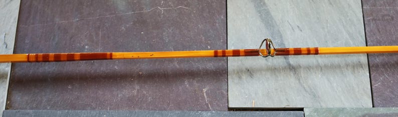 K&ouml;nnte beinhalten: Eine Angelrute aus Holz mit braunem und orangefarbenem Streifenmuster. Die Rute hat einen Metallring am Ende.