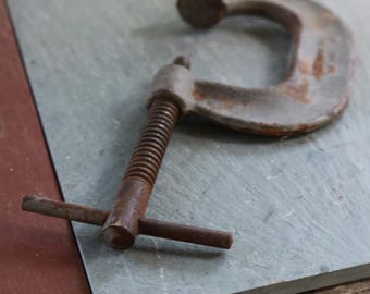 May include: A rusty C-clamp tool with a threaded rod and handle, resting on a gray stone tile. The clamp is made of metal and shows signs of wear. The background includes a red tile and a wooden surface.