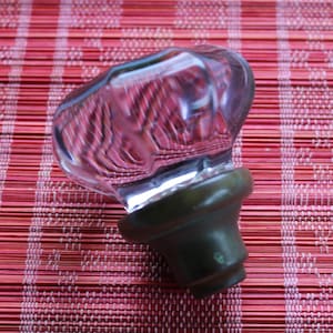 Könnte beinhalten: Ein klarer Glasflaschenverschluss mit abgerundeter Oberseite und dunkelgrüner Basis. Der Verschluss reflektiert das rot-weiße Muster der Oberfläche, auf der er ruht. Der Verschluss ist für eine Flasche konzipiert.